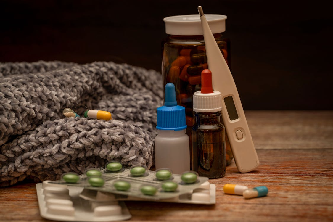 The image shows a collection of medical items, including pills, a thermometer, eye drops, and a blanket, likely indicating a person is unwell.