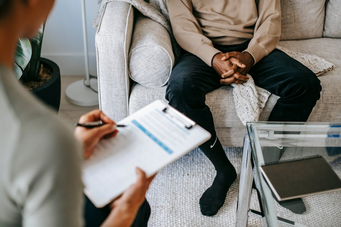 The image shows a therapist or counselor taking notes while a patient sits on a couch, possibly during a therapy session.