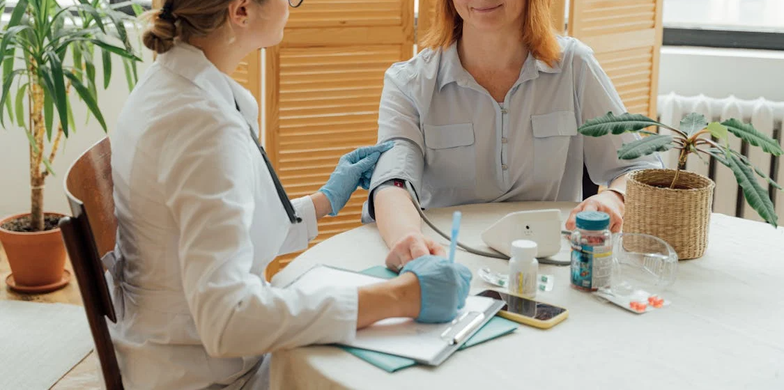 The image shows a healthcare professional in a white coat taking a blood sample from a patient, who is sitting and smiling, in a calm and well-lit environment.