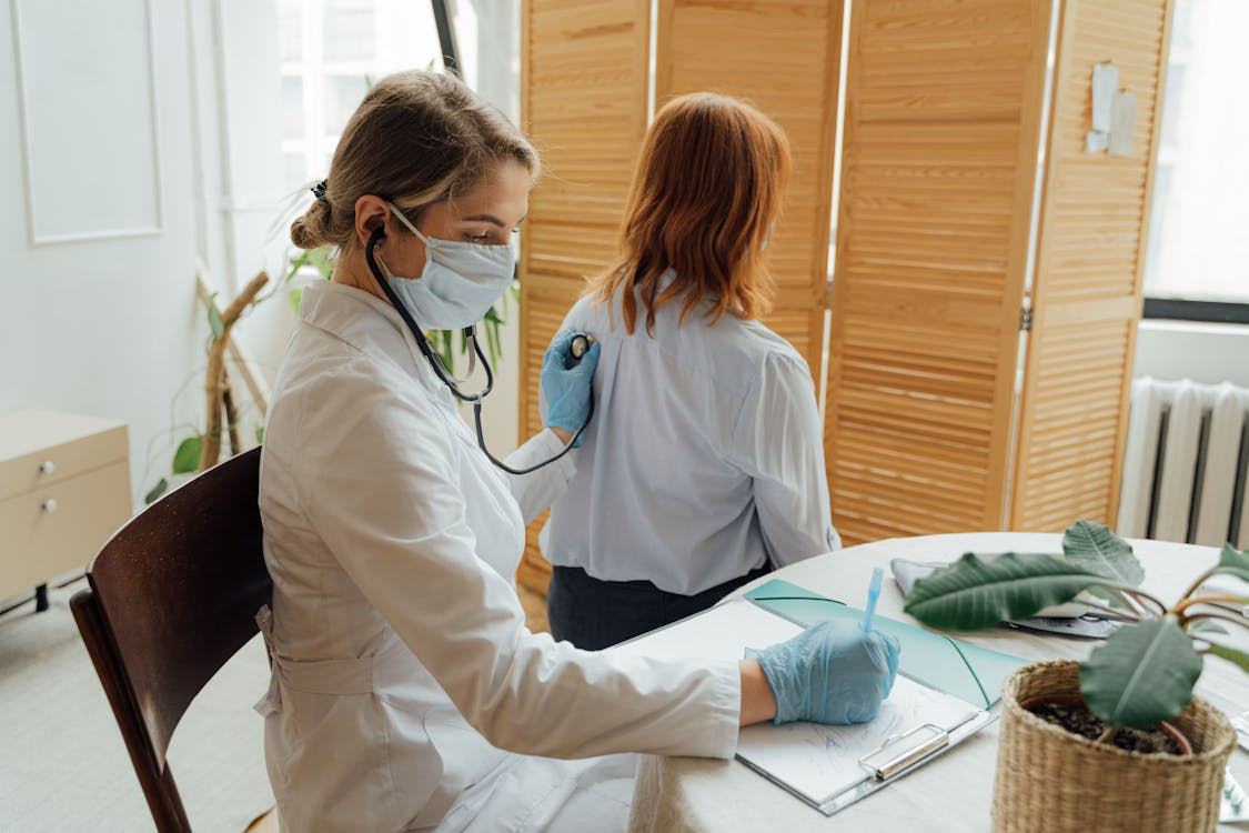 The image shows a healthcare professional listening to a patient's back with a stethoscope while taking notes during a check-up.