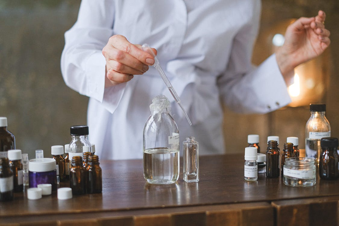 The image shows a person in a lab coat experimenting. They are using a pipette to transfer a liquid from one container to another, surrounded by various bottles and jars.