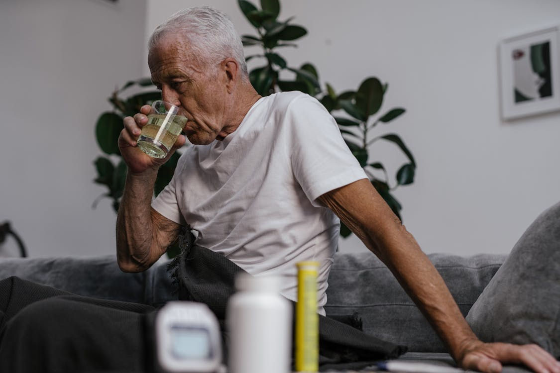 Elderly man drinking water from a glass with pill bottles in front of him